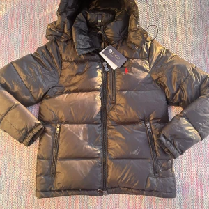 Polo Raulph Lauren jacka - ✔ Brand: Polo Ralph Lauren ✔ Model: Puffer Jacket ✔ Color: Glossy Black ✔ Size: Small (S) ✔ Condition: Brand new, worn as a trpohy ✔ Material: High-quality, water-resistant fabric with a sleek, glossy finish ✔ Features: Full-zip closure with a high collar for extra protection against the cold Elasticated cuffs and hem to retain warmth Two zippered side pockets for convenience and security ✔ Fit: Regular fit – warm yet lightweight, perfect for layering  A stylish and functional winter essential, 