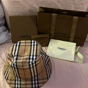 Rutig bucket hat från Burberry - Säljer en klassisk rutig bucket hat från Burberry i beige, svart och röd. Hatten är tillverkad i 100% bomull och har det ikoniska Burberry-mönstret. Perfekt för att ge en stilren touch till din outfit.