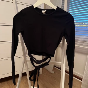Svart långärmad topp med knytband - Snygg svart långärmad topp med knytband i midjan. Perfekt för en stilren look. Toppen är croppad och har en enkel design som passar till många olika outfits.