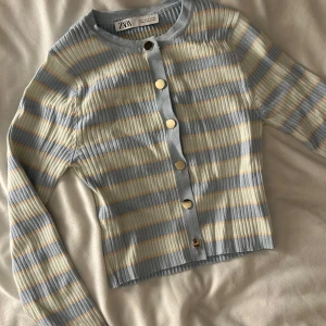 Randig ribbad kofta från Zara - Säljer en snygg randig kofta från Zara i ljusblått och beige. Den är ribbad med långa ärmar och har guldiga knappar framtill. Storlek M men passar allt från XS-M. Aldrig använd!