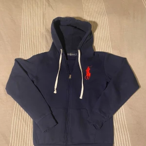 Hoodie från Ralph Lauren - Säljer en snygg mörkblå hoodie från Ralph Lauren med dragkedja och en stor röd broderad logga på bröstet. Tröjan är tillverkad i 100% bomull och har en justerbar huva med vita snören. Perfekt för en avslappnad stil.