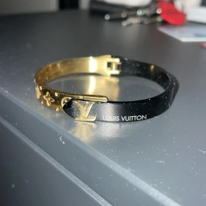 Guld och svart armband från Louis Vuitton - Elegant armband i guld och svart från Louis Vuitton. Armbandet har det ikoniska LV-märket och ett mönster med små symboler. Perfekt för att ge en lyxig touch till din stil. Tillkommer inget og, bara armbandet. Kontakta mig innan köp!