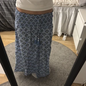 Blå mönstrad långkjol - Säljer en blå långkjol med ett snyggt mönster i vitt och mörkblått. Kjolen har en elastisk midja med knytband och tofsar. Går inte att köpa på Gina längre köpt från i somras.