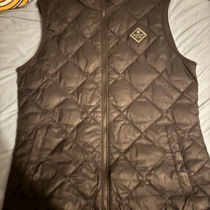 Svart quiltad väst från Gant - Snygg svart quiltad väst från Gant med dragkedja framtill. Perfekt för lager-på-lager under kyligare dagar. Klassisk design med en stilren logotyp på bröstet.