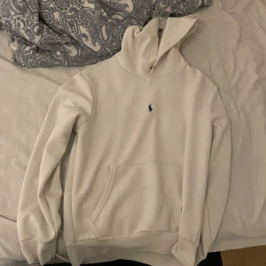 Vit hoodie från Ralph Lauren - Säljer en stilren vit hoodie från Ralph Lauren med det klassiska logotypbroderiet på bröstet. Tröjan har en känguruficka och en bekväm huva. Perfekt för en avslappnad look.
