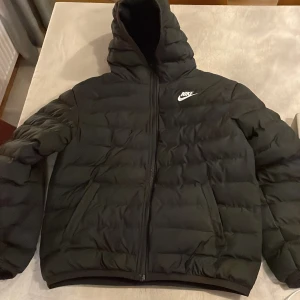 Svart dunjacka från Nike - Säljer en svart dunjacka från Nike med huva och dragkedja. Jackan har en puffig design och är perfekt för kyligare väder. Den har en klassisk Nike-logga på bröstet..