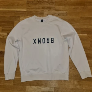 Crewneck sweatshirt - Säljer en stilren beige sweatshirt från SOC med texten 'BRONX' i svart på framsidan. Tröjan är tillverkad i en mjuk bomullsblandning och har långa ärmar samt rund halsringning. Perfekt för en avslappnad look. Skicket är 10/10