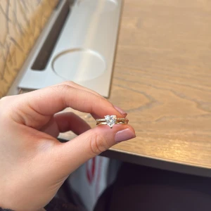 Guldfärgad ring med hjärtformad sten - Elegant guldfärgad ring med en stor hjärtformad klar sten i mitten. Ringen har en glittrig design med små stenar längs bandet som ger extra bling. Perfekt för speciella tillfällen eller som en vacker accessoar till vardags. Detta är en pandora ring som använts i tre månader bara och du kan även få med pandora boxen! 