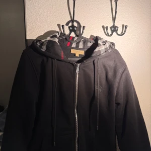 Svart huvtröja från Burberry - Snygg svart huvtröja från Burberry med dragkedja och klassiskt rutigt mönster i huvan. Perfekt för en stilren look med en touch av lyx. Tröjan har långa ärmar och en bekväm passform.