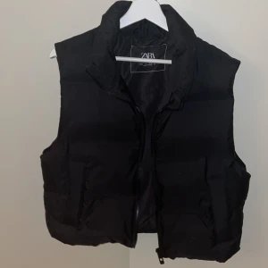Väst ifrån Zara - Snygg svart dunväst från Zara med dragkedja framtill. Perfekt för lager-på-lager under kyligare dagar. Västen har en hög krage och två praktiska fickor.