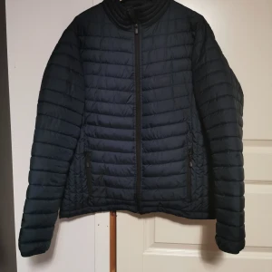 Svart pufferjacka från Tee Jays - Säljer en svart pufferjacka från Tee Jays. Jackan har en dragkedja framtill och är tillverkad i ett lätt och vattenavvisande material. Perfekt för kyligare dagar med sin quiltade design och långa ärmar.