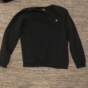Svart tröja från Lyle & Scott Junior - Säljer en stilren svart sweatshirt från Lyle & Scott storlek s med ett gult broderat emblem på bröstet. Tröjan har långa ärmar och rund halsringning. Perfekt för en avslappnad look. Skick 9/10 pris 300
