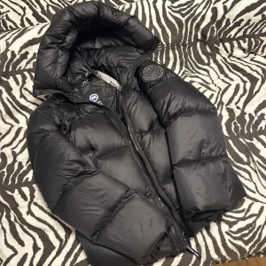 Svart Canada Goose pufferjacka  - Snygg svart pufferjacka med huva och dragkedja. Perfekt för kalla dagar med sin quiltade design som håller värmen. Jackan har en stilren look och passar bra till olika outfits.
