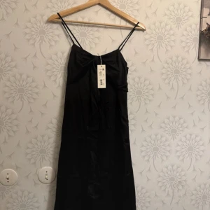 Svart axelbandslös klänning JC - Elegant svart klänning med smala axelband och en dekorativ rosett framtill. Perfekt för festliga tillfällen.