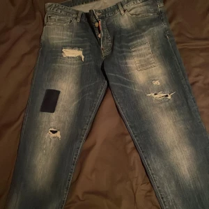 Blå jeans från Dsquared2 - Säljer ett par blå jeans från Dsquared2 med slitna detaljer och patchar. Jeansen har en broderad lönnlöv på bakfickan och knappar i gylfen. Perfekta för en avslappnad stil. Knappt använda