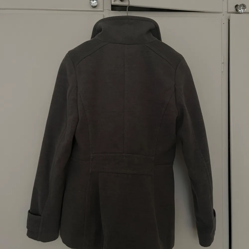 Snygg grå kappa med dubbelknäppning och stilren design. Jackan har långa ärmar och en klassisk krage. . Takit.
