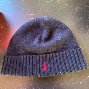 Mörkblå mössa från Polo Ralph Lauren - Snygg mörkblå mössa från Polo Ralph Lauren med röd broderad logotyp framtill. Tillverkad i mjukt material för extra komfort. Perfekt för kyliga dagar. Helt ny aldrig använd