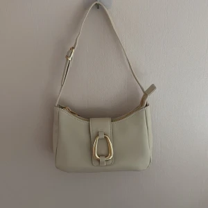 Beige axelväska med gulddetalj - beige axelväska med justerbar rem och gulddetaljer. Aldrig använd innan, nypris 300kr