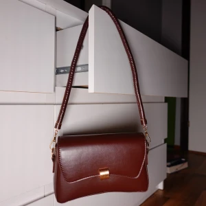 Kaffebrun axelväska med gulddetalj - Snygg brun axelväska i skinn med en elegant gulddetalj på framsidan. Väskan har en justerbar axelrem och en stilren design som passar perfekt för olika tillfällen.