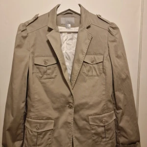 Beige kavaj från H&M - Snygg beige kavaj från H&M i lite tuffare stil. Perfekt när man vill se stilfull ut och dessutom sticka ut. Använd vid 2 tillfällen endast!  Garderobsrensning så kika gärna in övriga auktioner.