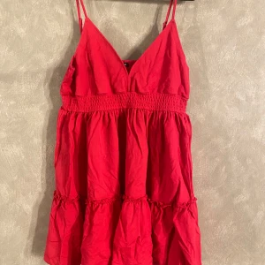 Röd klänning från Sinsay - Snygg mörkrosa klänning från Sinsay med smala axelband och v-ringad design. Klänningen har en luftig passform med volanger och resår i midjan för en bekväm känsla. Bilden uppfattas som röd, men är tydligt mörk rosa i verkligheten. Står att det är en XL men passar mig som är S/M☺️