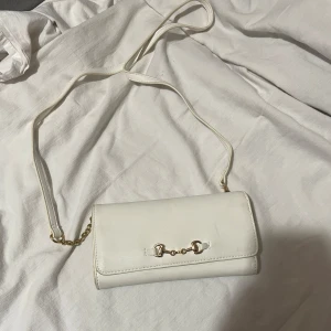 Vit axelväska med gulddetaljer från Glitter  - Snygg vit axelväska i syntetmaterial med gulddetaljer. Väskan har en elegant kedja och en praktisk axelrem. Perfekt för att bära det viktigaste med stil.  Haft den länge men den är ftf fint väska!
