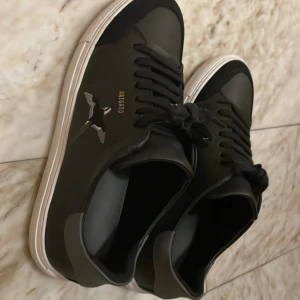 Svarta sneakers från Axel Arigato - Snygga svarta sneakers från Axel Arigato med vit sula och gulddetaljer. Skorna har en stilren design med snörning och en diskret fågelbrodering på sidan.