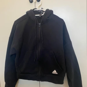 Svart hoodie från Adidas - Säljer en svart hoodie från Adidas med klassiska vita ränder längs ärmarna. Den har dragkedja och en bekväm huva. Bara använd några gånger. Aningen liten i storleken