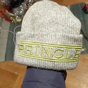 Grå ribbad mössa från Pringle - Snygg grå ribbad mössa från Pringle med broderad logga i gult. Perfekt för att hålla värmen under kyliga dagar. Stilren design som passar till det mesta.