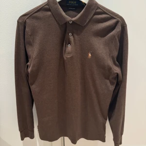 Brun pikétröja från Polo Ralph Lauren - Säljer en stilren brun pikétröja från Ralph Lauren i pimabomull. Tröjan har långa ärmar och en klassisk krage med knappar framtill. Perfekt för en avslappnad men ändå elegant look. Custom slim fit. Nypris 1500kr