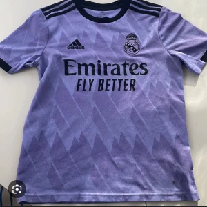 Lila fotbollströja från Adidas ( storlek Xs och S - Snygg lila fotbollströja från Adidas med Real Madrid-logga och 'Emirates Fly Better' tryck på framsidan. Tröjan har korta ärmar och ett diskret mönster i tyget. Perfekt för fotbollsfans!