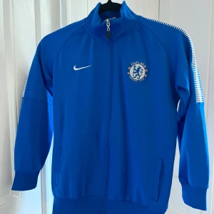 Chelsea träningsjacka från Nike - Säljer en blå Chelsea jacka från Nike med dragkedja och klubbens emblem på bröstet. Jackan har långa ärmar och ribbade muddar. Perfekt för fotbollsfans! Juniorstorlek  L - 164 (obs liten i storlek) Jackans rygglängd 58 cm, bredd 45 cm. Ärmlängd ca 45 cm från axel.
