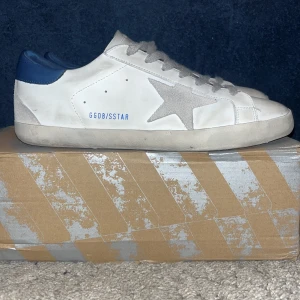 Vita sneakers med blå detaljer från Golden Goose - Snygga vita sneakers från GGDB med en grå stjärndetalj på sidan och blå häl. Skorna har grå snörning och en klassisk design som passar perfekt till vardags. De är tillverkade i skinn och har en bekväm passform. Skiktet är 6/10 , om mer bilder önskans så kom privat.