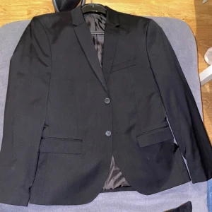 Svart kavaj från H&M - Snygg svart kavaj från H&M med två knappar framtill och klassiska slag. Perfekt för en stilren look. Kavajen har fickor med lock och en innerficka för extra förvaring.