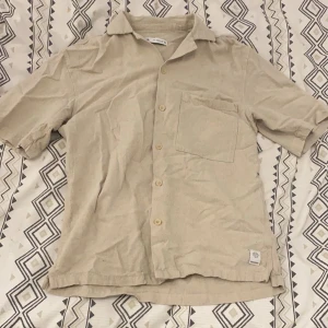 Beige kortärmad linneskjorta - Säljer en stilren beige kortärmad skjorta med knappar och en bröstficka. Perfekt för varma dagar och avslappnade tillfällen.