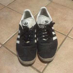 Mörkblåa Adidas Gazelle sneakers - Säljer ett par klassiska mörkblåa Adidas Gazelle sneakers med vita detaljer och snörning. Skorna har den ikoniska tre-randiga designen och en bekväm passform.