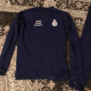 Mörkblå träningsset från Global Football Barcelona - Säljer ett mörkblått träningsset från Global Football Barcelona. Tröjan har långa ärmar och en halv dragkedja vid halsen. Byxorna har en elastisk midja och logotyp på benet. Perfekt för fotbollsträning eller annan sportaktivitet.