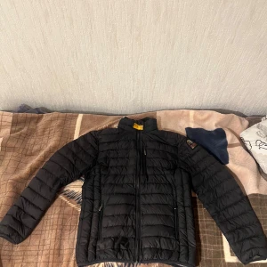 Svart dunjacka med dragkedja - Snygg svart dunjacka från Parajumpers med dragkedja och hög krage. Jackan har en quiltad design och är perfekt för kyligare väder. Insidan är grå och ger en fin kontrast till den svarta utsidan. passa s-l