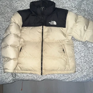  The North Face retro nuptse puffer - Säljer en beige och svart dunjacka från The North Face. Jackan har en dragkedja framtill och är långärmad. Perfekt för kyliga dagar med sin pufferdesign som håller dig varm. Har några fläckar på sig som definitivt försvinner med en tvätt. 