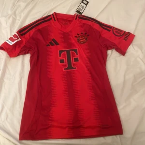 Röd FC Bayern München fotbollströja från Adidas - Säljer en snygg röd FC Bayern München fotbollströja från Adidas. Tröjan har korta ärmar och är i nyskick med alla originaltaggar kvar. Har aldrig använt! Öppen för pris ändring, i så fall skriv till mig.