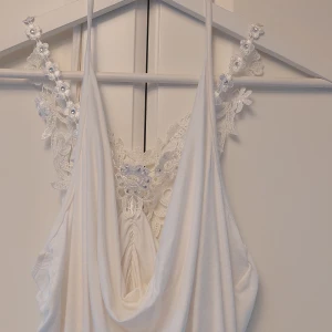 Vit topp med spetsdetaljer - Säljer en superfin vit topp med vackra spetsdetaljer och gnistrande stenar.. Toppen har en draperad framsida och smala axelband. Den är i nyskick och passar perfekt till både jeans och kjol. Köpt i Italien. Storleksmärkt som M, men är mer S.