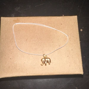 Armband med elefantberlock - Säljer ett enkelt och stilrent armband med en söt elefantberlock i guld. Armbandet har en tunn vit tråd som ger en minimalistisk look. Perfekt för vardagsbruk eller som en fin present!