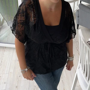 Svart spetsblus med knytband - Säljer en svart spetsblus med knytband i midjan. Blusen har korta ärmar och är omlott. Supersnygga detaljer. Är i Storlek L men går också att justera att göra mindre💞
