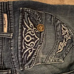 Säljer ett par snygga blå jeans med coola broderade detaljer på bakfickorna. Jeansen har en klassisk femficksdesign och är i en skön denimkvalitet. Perfekta för en avslappnad vardagslook!