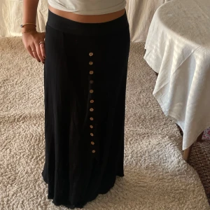 Lång svart kjol  - Ribbad svart långkjol, bra skick, knappar och en slits nedanför 