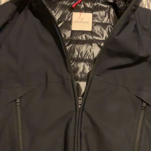 Svart jacka från Moncler - Säljer en snygg svart jacka från Moncler. Den har en stilren design med dragkedja och Moncler-logga på ärmen. Jackan är perfekt för kyligare väder och har en quiltad insida för extra värme. Passar både till vardags och fest!