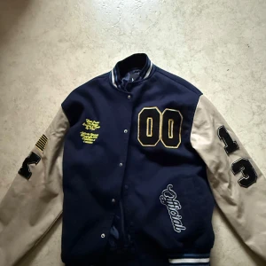 Blå och beige bomberjacka från BoohooMAN - Snygg bomberjacka i blått och beige från BoohooMAN. Jackan har långa ärmar med nummerdetaljer och broderade märken. Den har en klassisk college-stil med knappar framtill och ribbade muddar vid ärmslut och nederkant.