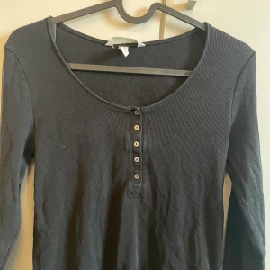 Svart ribbad långärmad topp - Säljer en svart ribbad långärmad topp med knappar framtill. Perfekt för en stilren och bekväm look. Passar bra till både jeans och kjol.