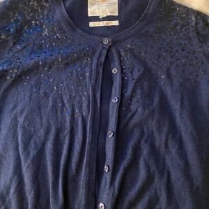 Mörkblå kofta  - Säljer en snygg mörkblå kofta från Holly & Whyte by Lindex. Koftan har långa ärmar och är dekorerad med glittriga paljetter på axlarna. Perfekt för att lägga till lite extra stil till din outfit. Knappar framtill för enkel stängning.