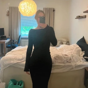 Svart långklänning med öppen rygg - Elegant svart långklänning med långärmad design och en stilfull öppen rygg. Perfekt för en kväll ute eller en speciell tillställning. Klänningen har en tight passform. Klänningen är i ett glittrigt material 